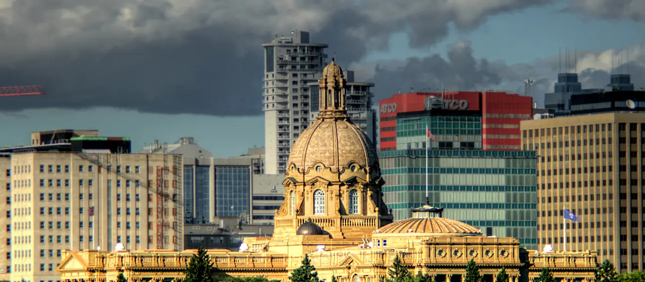 The domed Alberta Legislature Building lit by evening sun in front of downtown Edmonton office towers